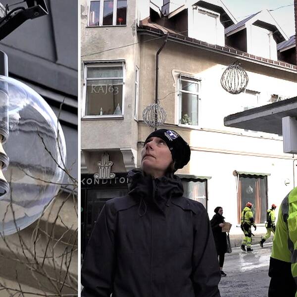 En kvinna med mörka kläder och en man med gulgrön jacka står i stadsmiljö och sneglar upp mot en gatlykta i form av en glasglob.