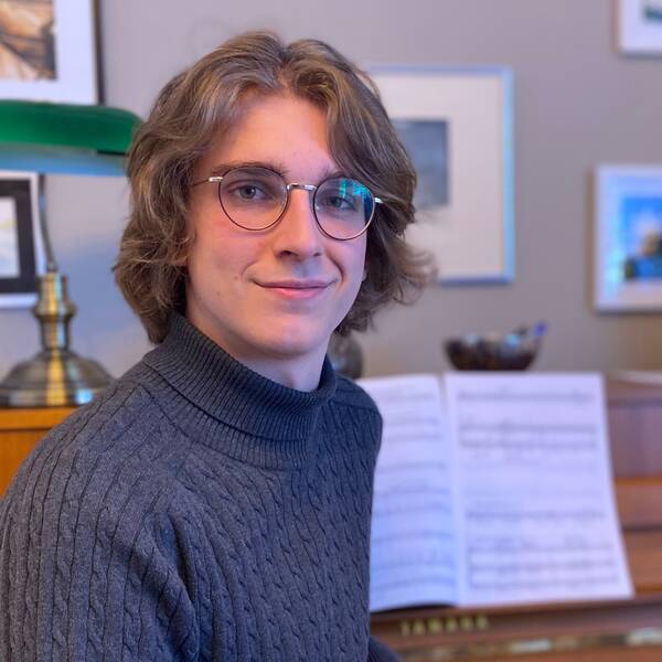 Milosz Krawczyk, en 19-årig kille med halvlångt hår och glasögon sitter framför ett piano med ett notblad och ler.