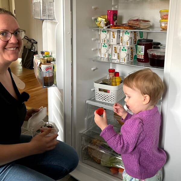 Klara Twete sitter på huk framför kylen och ger sin dotter en jordgubbe.