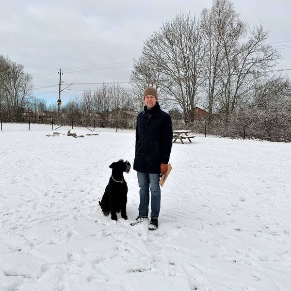 Sam Graflund Wallentin, en man i jacka och mössa, står ute i snön på en hundlekplats tillsammans med sin hund.