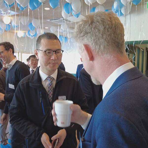 Vita och blå ballonger hänger taket. Flera personer står och pratar med varandra och ser glada ut. Donjings platschef Yun Lee lyssnar på en man som håller i en kaffekopp.