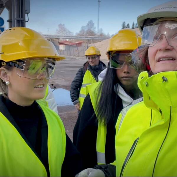 En kvinnlig ingenjör står omringad av unga tjejer i gula hjälmar och reflexvästar.