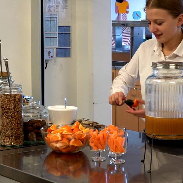 Flicka står i kök och skär en apelsin i klyftor. Har vit tröja på sig och brunt hår.