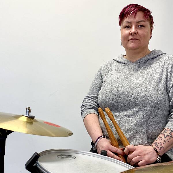 En bild på en kvinna i grå tröja, Emelie Eriksson. Hon sitter vid ett trumset och håller trumpinnar i handen.