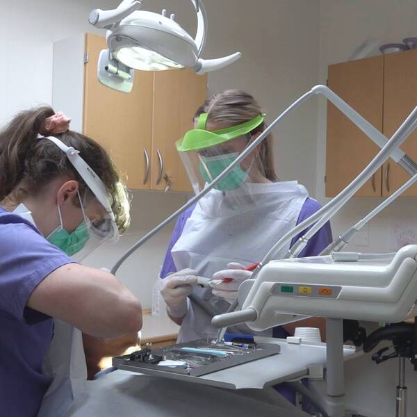 Två tandläkare står med munskydd och visir över en patient som får sina tänder undersökta.