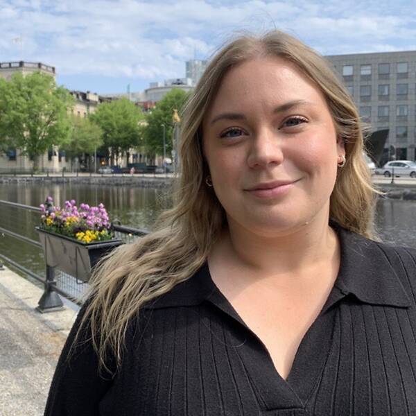 Tjej med halvlångt blont hår tittar in i kameran. I bakgrunden syns ån och stadshuset i Västerås.