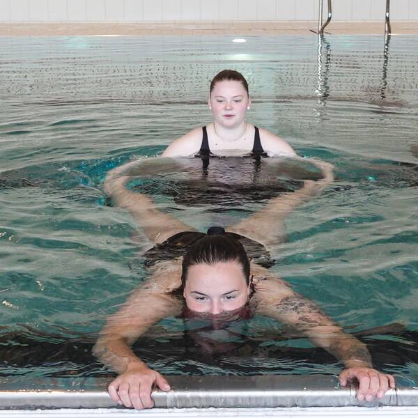 En bild på två tjejer, Victoria Lundqvist och Alice Tengelin, i en pool. Den ena av dem flyter på mage i vattnet och håller i poolkanten och den andra håller i hennes ben.