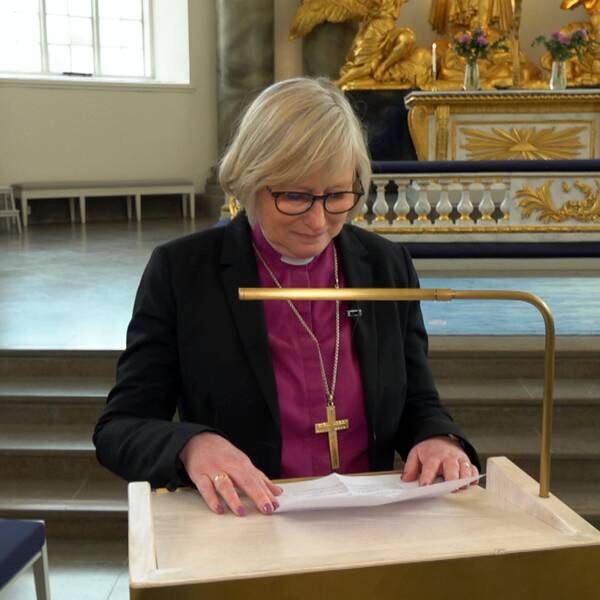 En bild på en kvinna, biskopen Susanne Rappmann, i rosalila skjorta, prästkrage, kavaj och korshalsband står vid en predikstol med ett papper i handen.