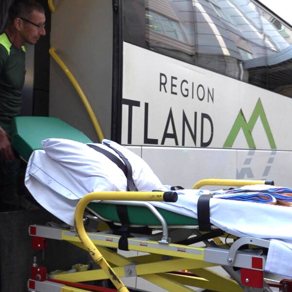 En ambulanssjukvårdare står i den bakre dörren av en vit buss med svart text. Han håller i en sjukhusbår och tittar ut åt sidan, bort från kameran.