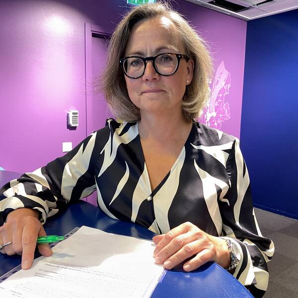 Kvinna står med ett papper framför sig. Hon har halvlångt brunt hår och glasögon. Tittar in i kameran.