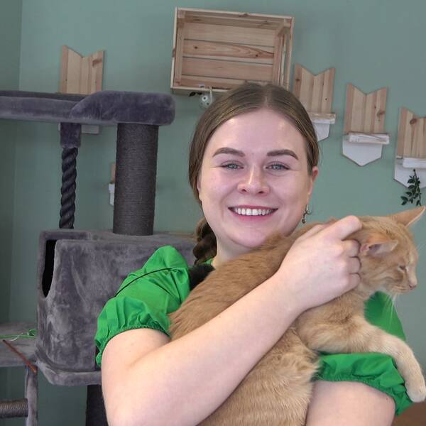 En kvinna ler och tittar rakt in i kameran. I famnen håller hon en röd katt som spinner. Hon har en grön tröja och i bakgrunden ses ett klätterträd för katter.