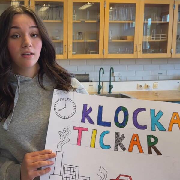 Amalia Lamnitchi står i ett klassrum på Gudmundråskolan i Kramfors. Hon har långt mörkt hår, grå huvtröja och håller i ett plakat med en klocka, industri och bil samt texten ”klockan tickar” i olika färger.