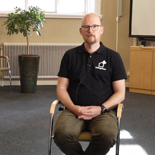 En man i bruna chinos, svart piké sitter på en stol i ett konferensrum. Han heter martin eneborg