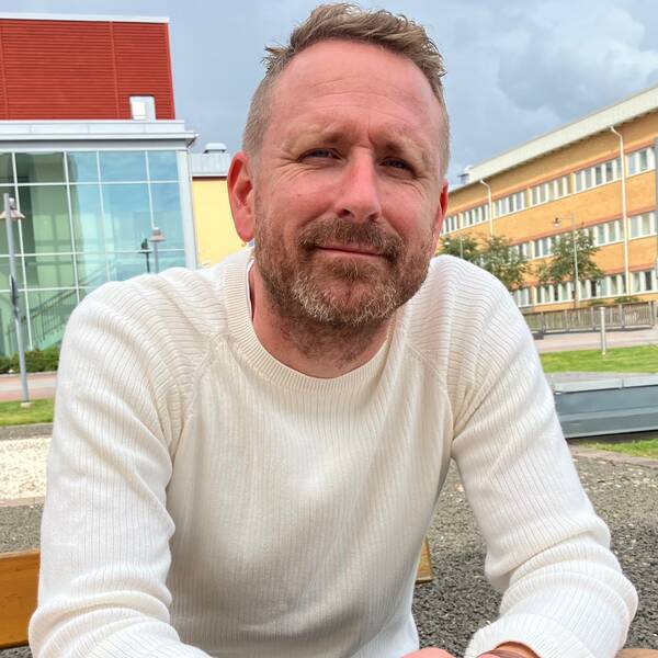 En man i vit tröja sitter på en bänk utomhus och tittar in mot kameran med ett litet leende.