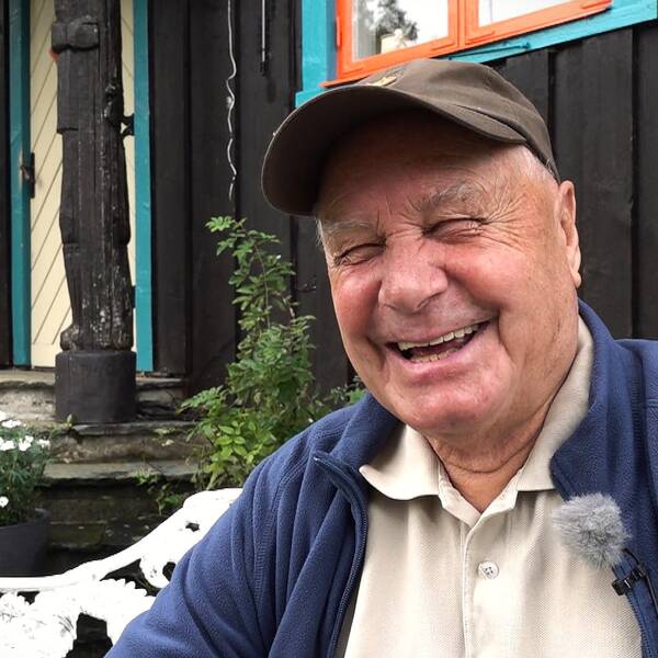 Glad äldre man i brun keps sitter framför en svartmålad träbyggnad med färgglada fönster.