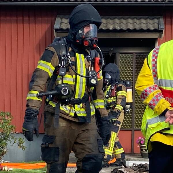 Rökdykare med svart huva och mask för ansiktet framför rött hus. Räddningsledare i gul jacka med röd hjälm står med ryggen mot kameran. Jämjö, Trädgårdsvägen, villabrand, övertänd.