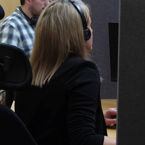 En svartklädd kvinna sitter med hörlurar framför en dator. Man ser henne snett bakifrån. Bakom henne sitter en man i rutig skjorta.