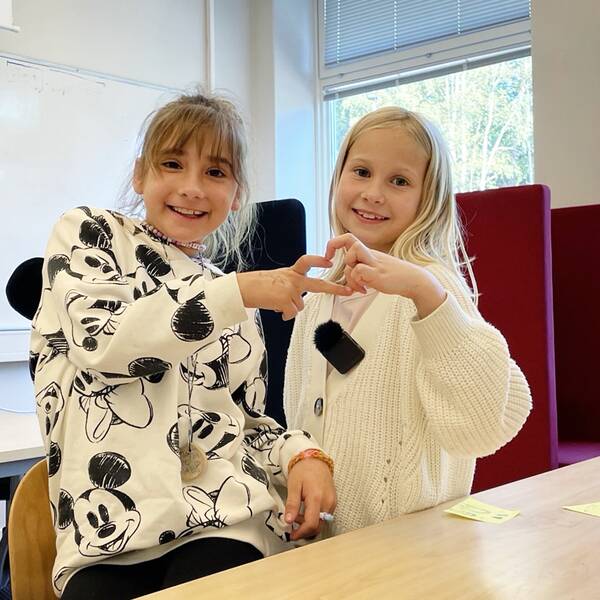 två 9-åriga flickor i vita tröjor, inne i ett klassrum håller fingrar mot varandra som bildar ett hjärta