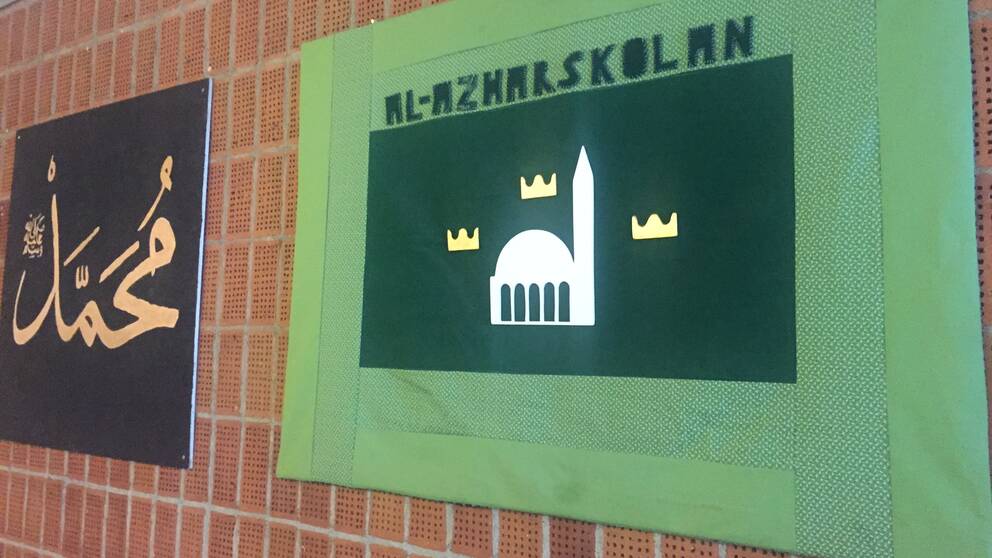 Två tygtavlor på tegelvägg. En svart med arabisk text på och en grön där det står Al-Azharskolan.