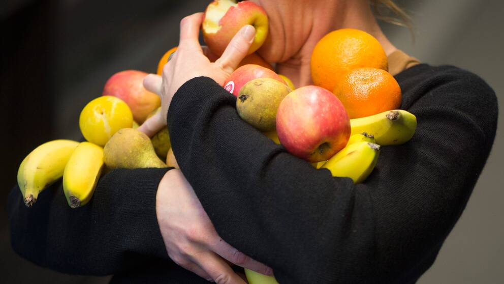 Kvinna med famnen full av frukt tar en tugga ur ett äpple