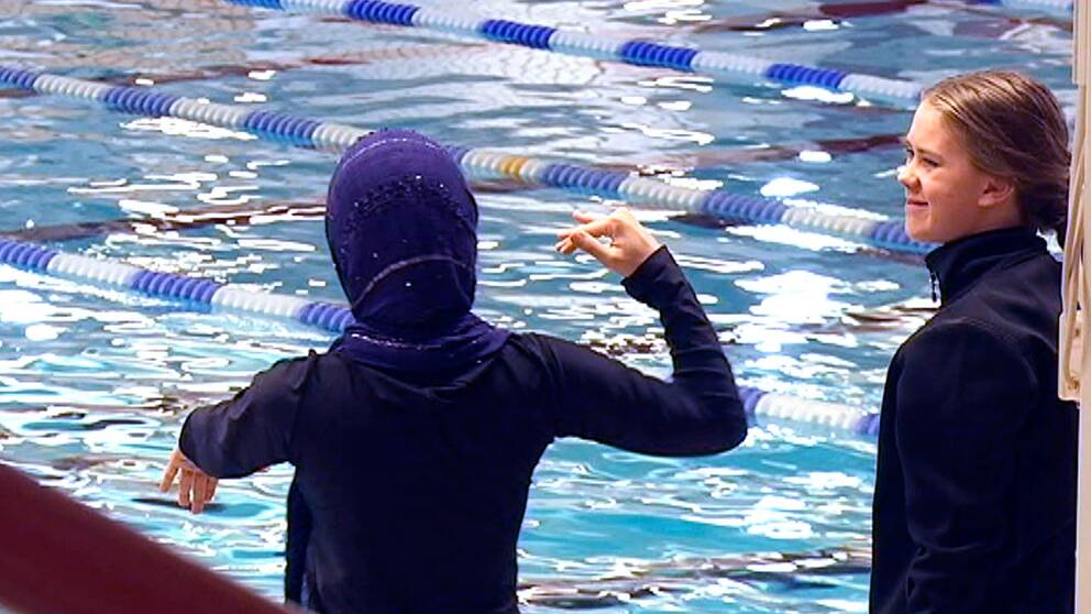 Flicka i burkini gestikulerar vid bassängkanten. Simhallspersonal ler.