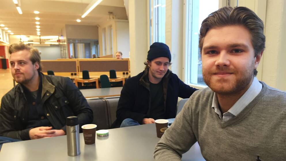 Tre studenter sitter vid ett bord. En ung man tittar in i kameran, han är till höger i bild.
