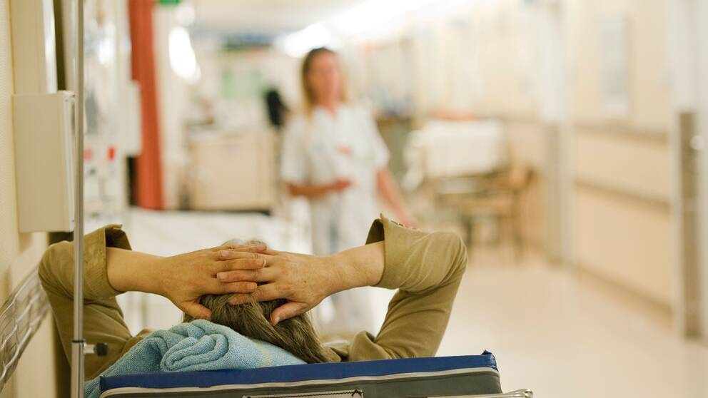 patient på akuten ligger i säng med händerna bakom huvudet, i bakgrunden en sjuksköterska i korridor