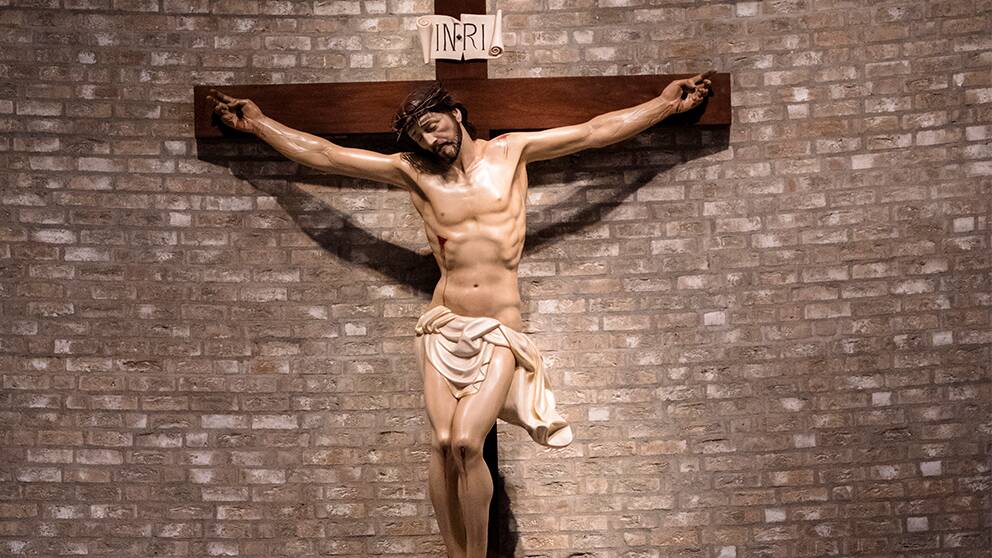 Bild på en staty vid en tegelvägg (förmodligen i en kyrka), där Jesus hänger på korset. Ovanför står det INRI på latin, vilket är en akronym för ”Jesus från Nasaret, judarnas konung”.