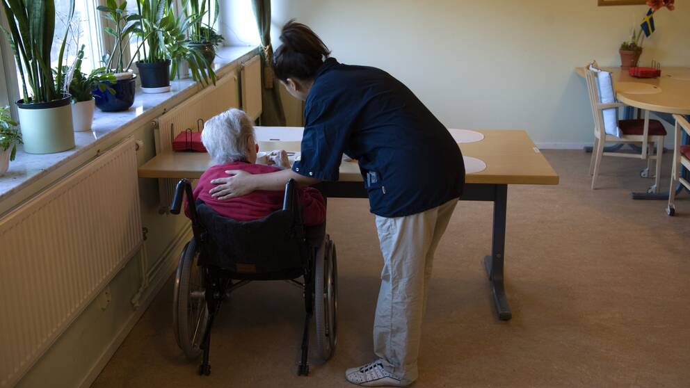 Arkivbild med äldre person i rullstol och sköterska ståendes bredvid, båda med ryggen mot kameran