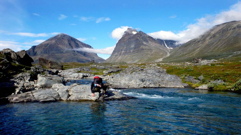 Fjällvandrare dricker vatten ur jokk. Fjäll i bakgrunden.