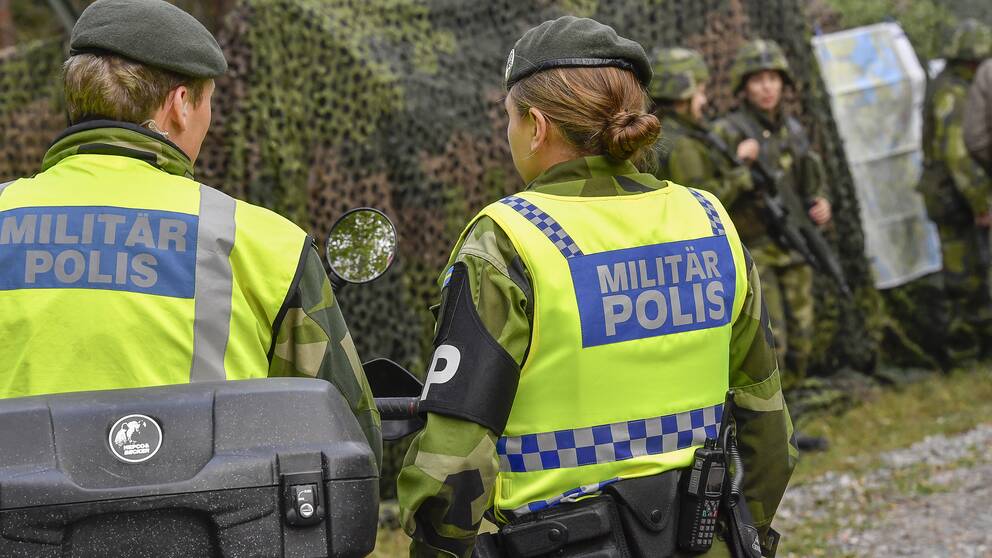 Två kvinnliga militärpoliser framför ett förband med soldater och ett kamoflerat militärfordon.