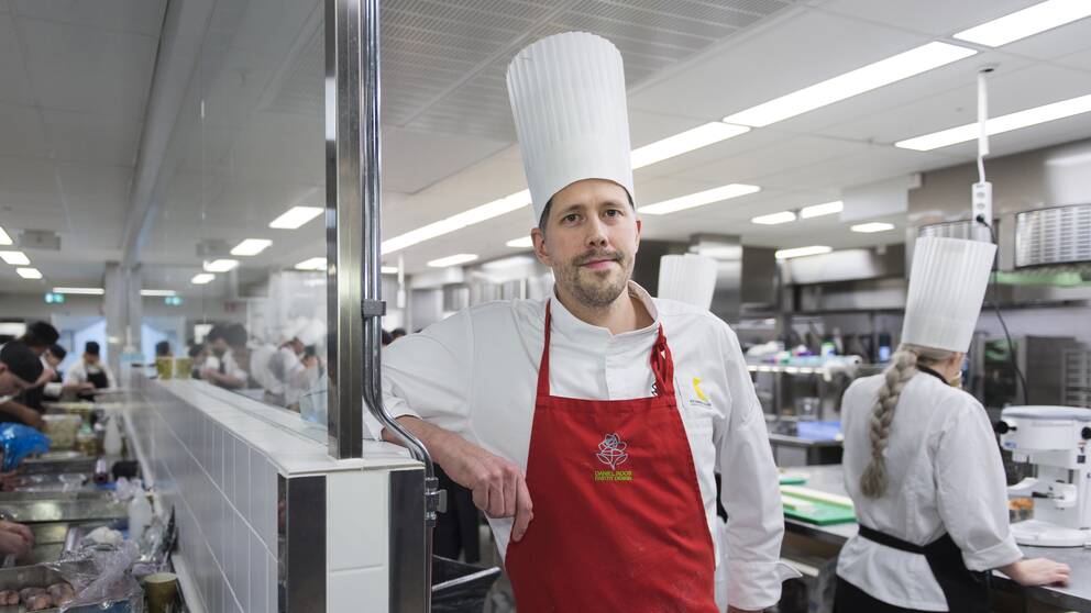 Daniel Roos står i ett kök med massa kockar som arbetar bakom. Han har på sig vit rockmössa, vit kockskjorta och rött förkläde.