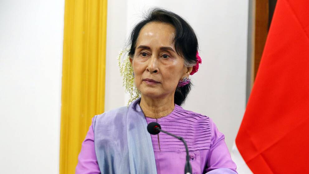 Aung San Suu Kyi står i ett talarpodium med blommor i håret.