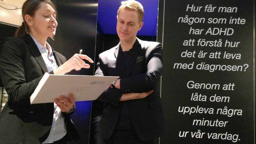 Forskaren pekar på ett test och berättar för utbildningsminister Gustav Fridolin som står med armarna i kors. Bakom dem finns adhd-lådan som har en öppen dörr och bredvid dörren står texten: ”Hur får man någon som inte har ADHD att förstå hur det är att leva med diagnosen? Genom att låta dem uppleva några minuter.”