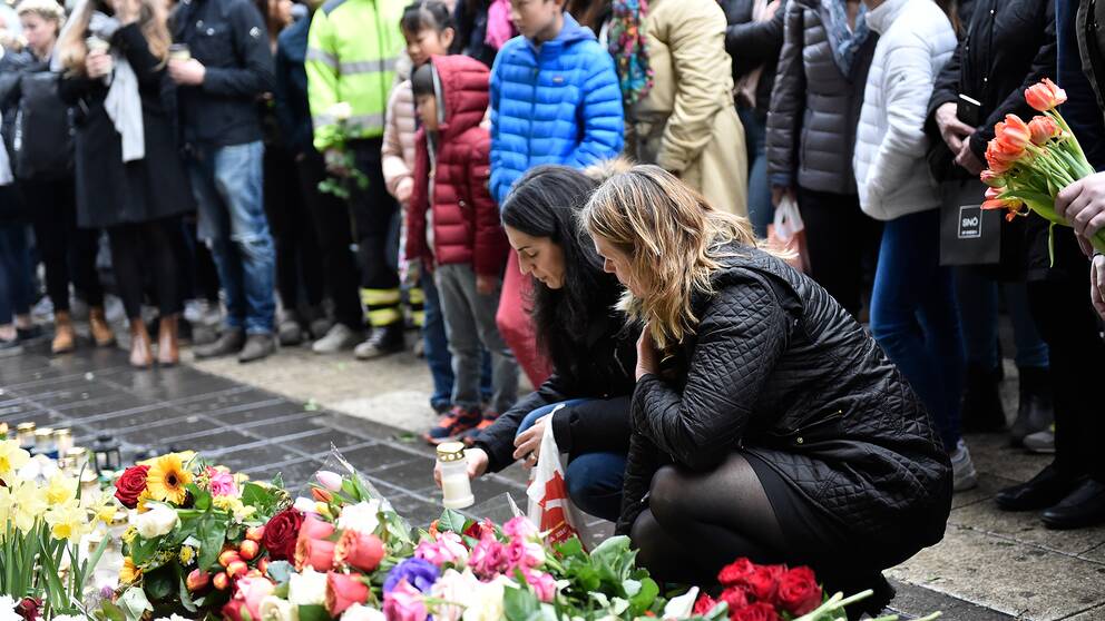 Två kvinnor står hukade framför ett blomsterhav för att hedra de som drabbades av terrorattacken på Drottninggatan i Stockholm 2017