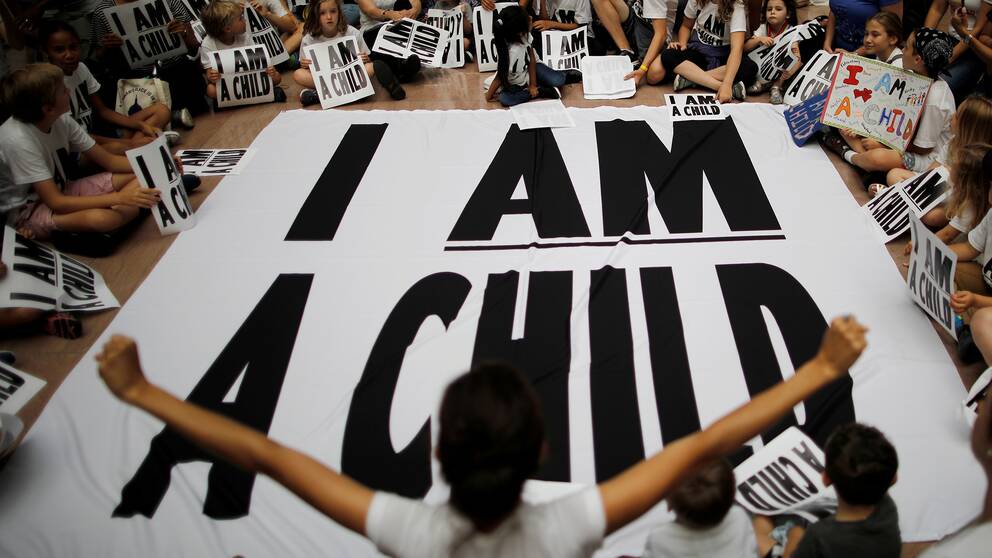 Barn sitter med plakat och en banderoll där det står ”Jag är ett barn”