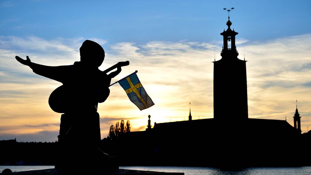 Evert Taube-statyn på Riddarholmen med en svensk flagga i famnen. Stadshuset i bakgrunden.