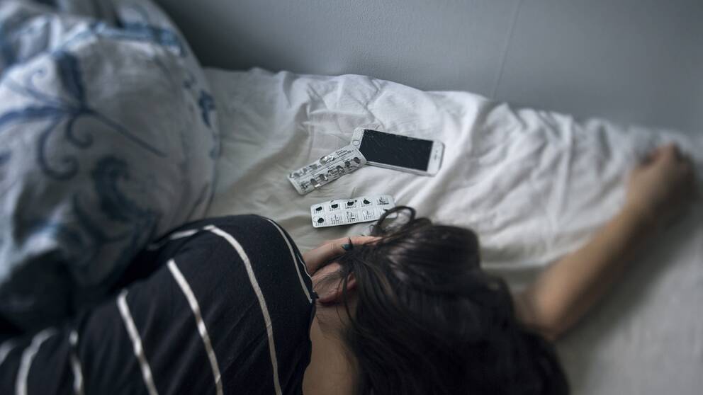 Ung kvinna ligger i sängen med tabletter och mobiltelefon intill huvudet.