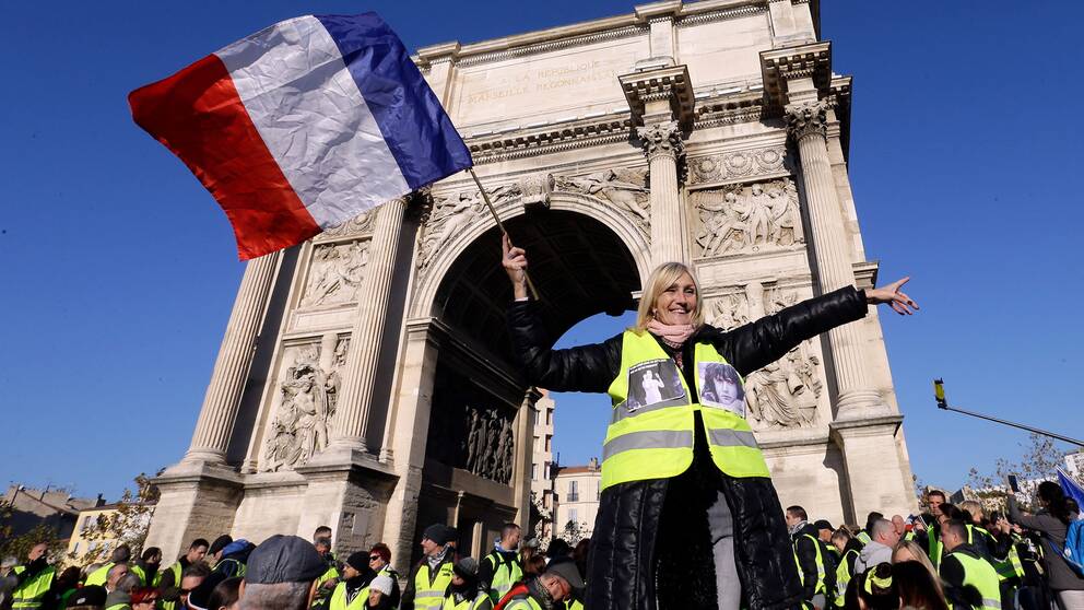 En demonstrant i gul väst viftar med den franska flaggan framför Triumfbågen i Marseille