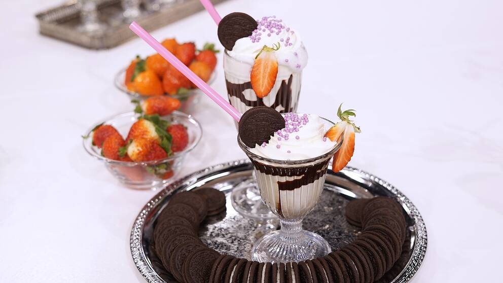 En metallbricka med två glas Mintchokladshake, dekorerade med chokladkex, strössel och jordgubbar. I bakgrunden finns två skålar med jordgubbar. 