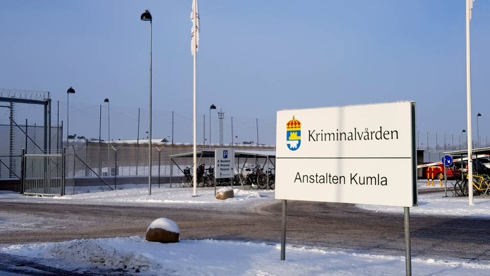 En skylt med orden Kriminalvården och Anstalten Kumla. Skylten står vid parkeringen utanför Kumlaanstalten, till vilken man ser stängslet i bakgrunden.