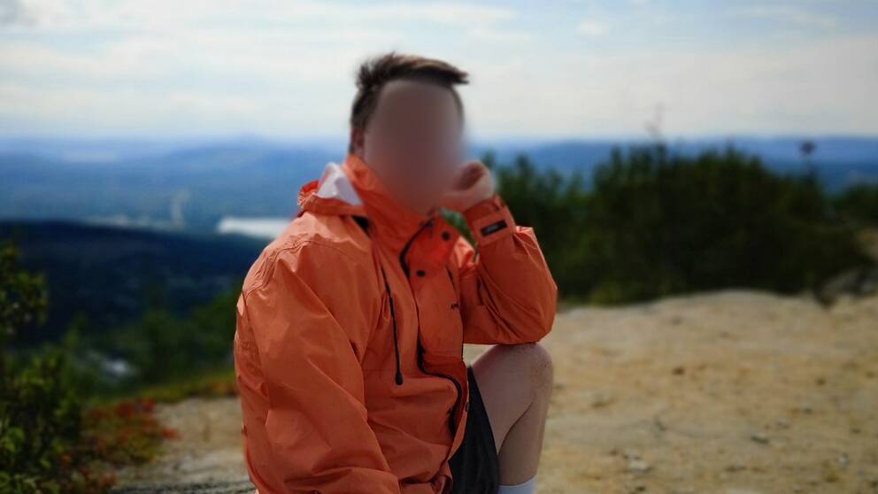 Man som sitter på en sten i naturen. Lutar huvudet i ena handen och har orange jacka.