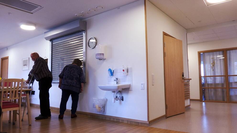 På bilden syns en vägg med en lucka där en gallerliknande jalusi har dragits ner. En kvinna tittar in genom gallret där det gemensamma avdelningsköket finns. 