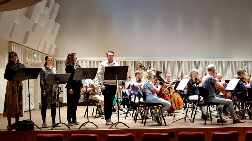 Till vänster i bild syns, från vänster, solisterna Hanna Wåhlin, Rebecca Fjällsby, Susanna Sundberg och Tobias Westman, alla framför varsitt notställ på scen. Till höger syns halva orkestern sittandes med instrument och blickarna vända mot höger.