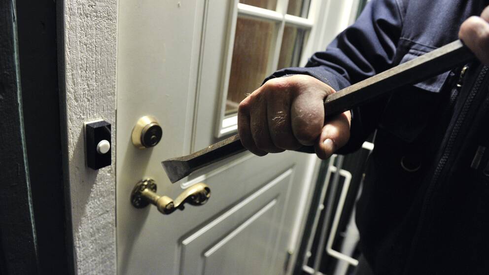 En man står vid dörren till en villa med en kofot i händerna, på väg att göra inbrott.