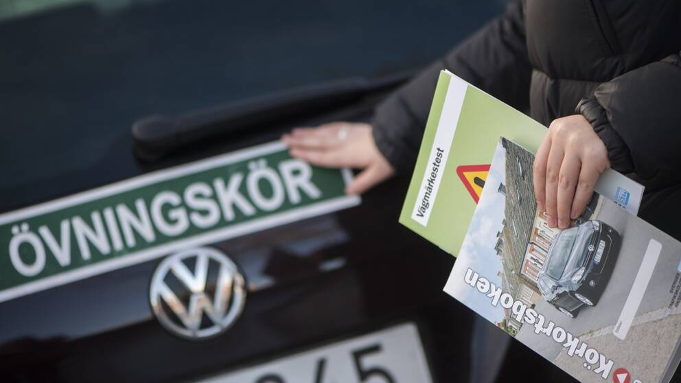 En person, som håller i körskoleböcker i handen, klistrar fast en övningskörningsskylt på en bil.