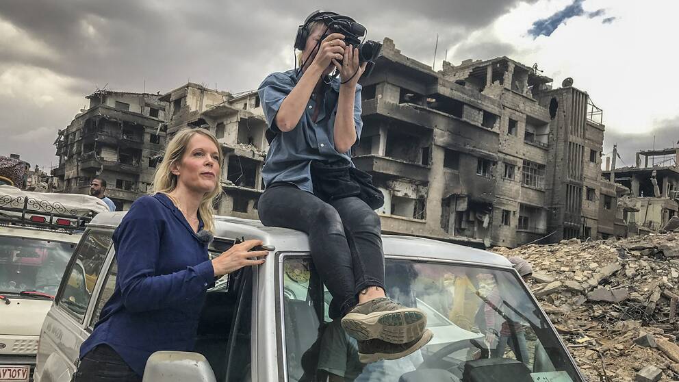Stina Blomgren och fotograf Pernilla Edholm i en krigshärjad stad. Pernilla sitter på taket på en bil.