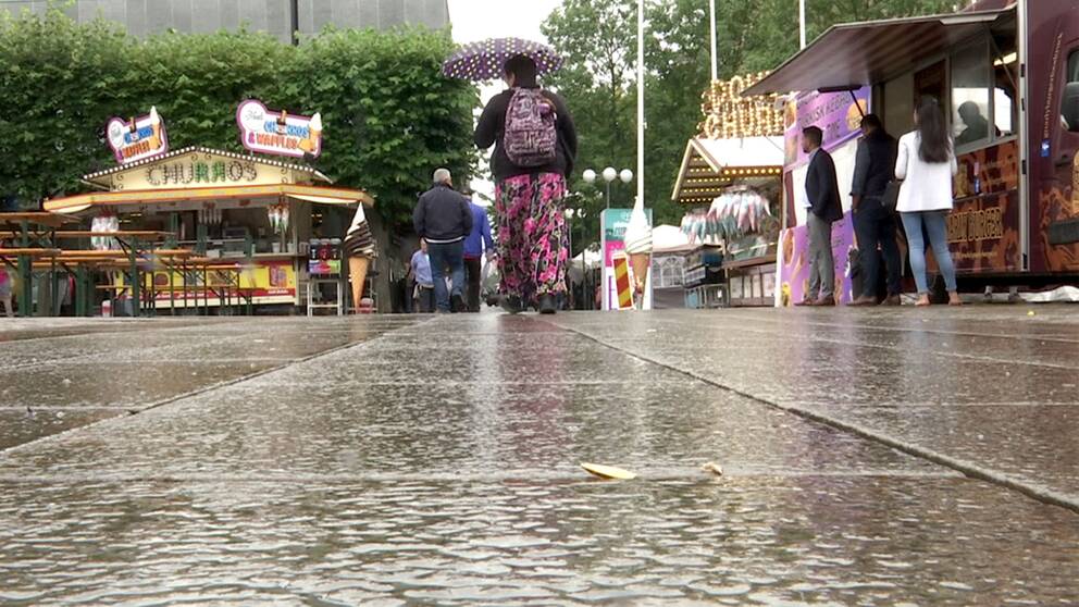Besökare på Södertäljefestivalen i regn och i mitten promenerar en dam med paraplymellan matstånden.