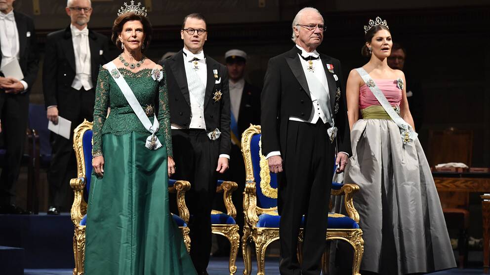 Drottning Silvia som bär på en grön klänning designad av Georg et Arend på nobelbanketten 2018. Snett bakom sig har hon svärsonen Prins Daniel och bredvid står Kung Carl XVI Gustaf. Längst till höger står kronprinsessan Victoria med en klänning som hyllades av kritikerna och som lär vara en homage till sin mor, som bar en liknande klänning på nobelgesten 1995.