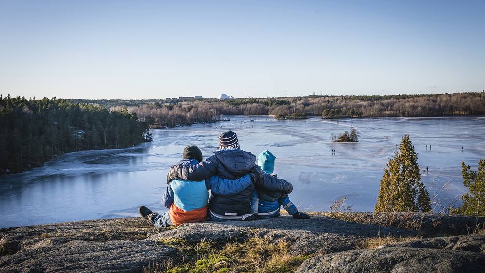 Tre personer sitter på klippa med utsikt över sjö.